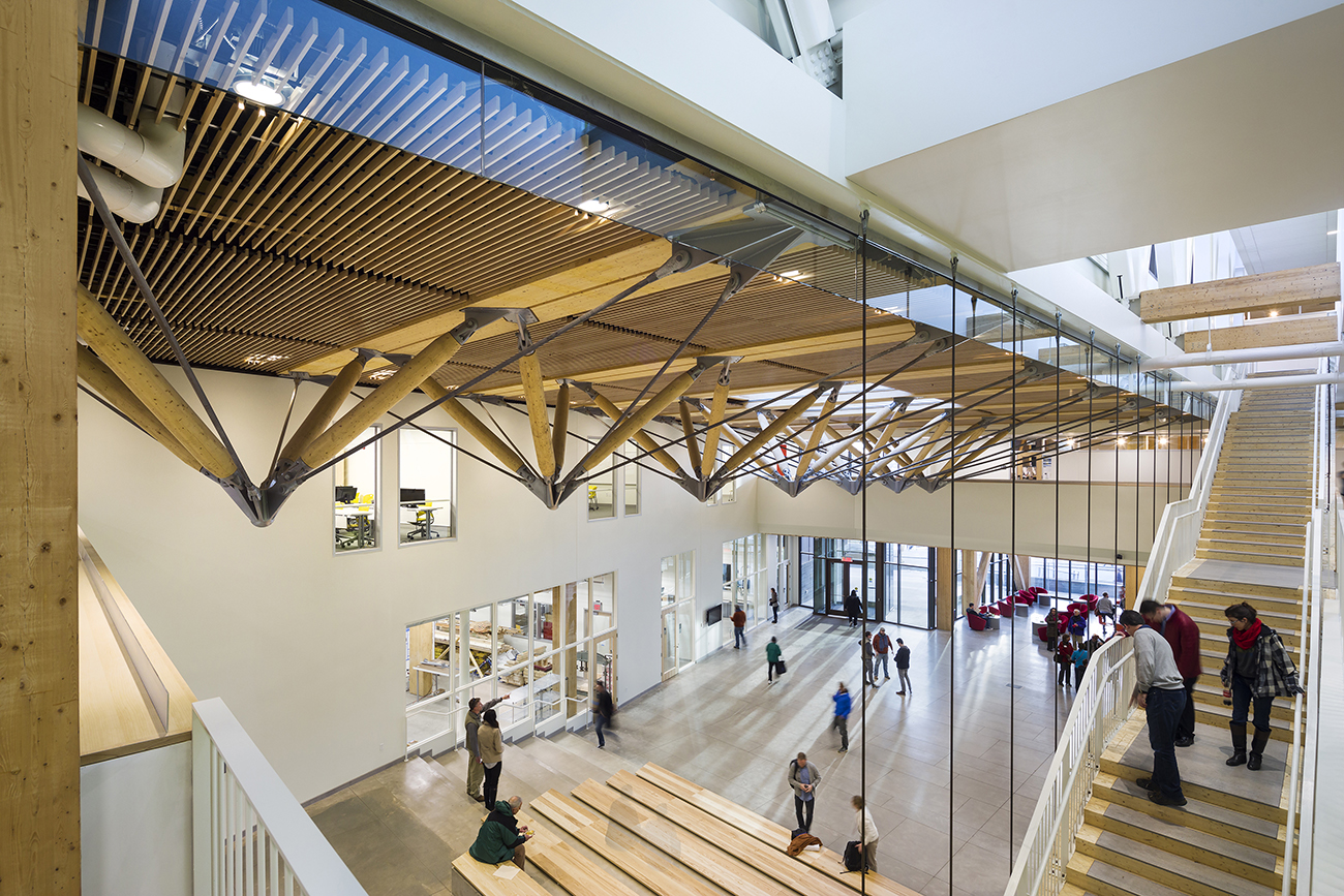 A modern, spacious building interior with exposed wooden beams, large windows, glass walls, and an open staircase. People are walking and conversing in the bright, airy atrium with natural light.