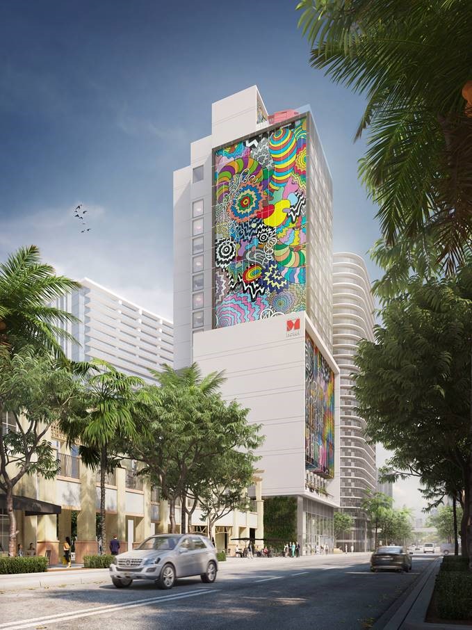 A tall building in a city with colorful, abstract murals on its facade; cars drive by on a palm tree-lined street under a partly cloudy sky.