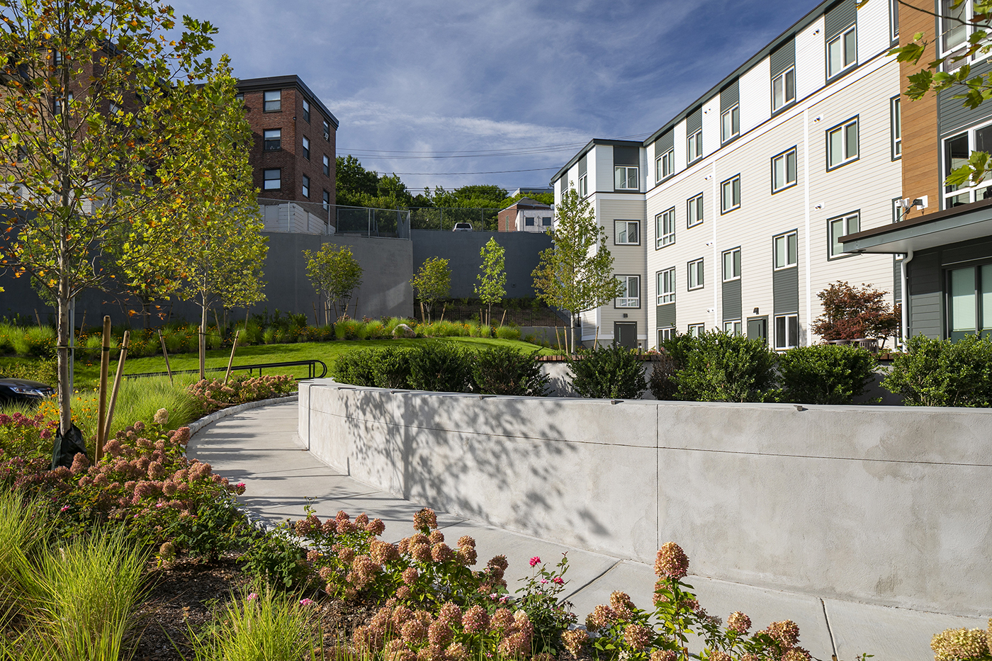 A winding concrete pathway curves through landscaped gardens with shrubs and young trees, leading to a modern, multi-story apartment building under a blue sky.