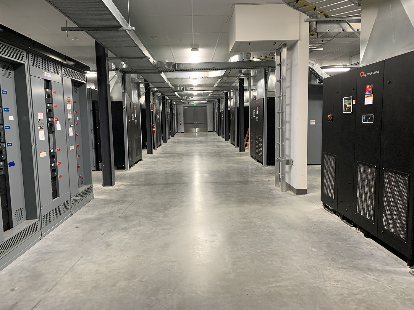 A wide, empty hallway in a data center with rows of server racks and electrical panels on both sides, bright overhead lighting, and a polished concrete floor.