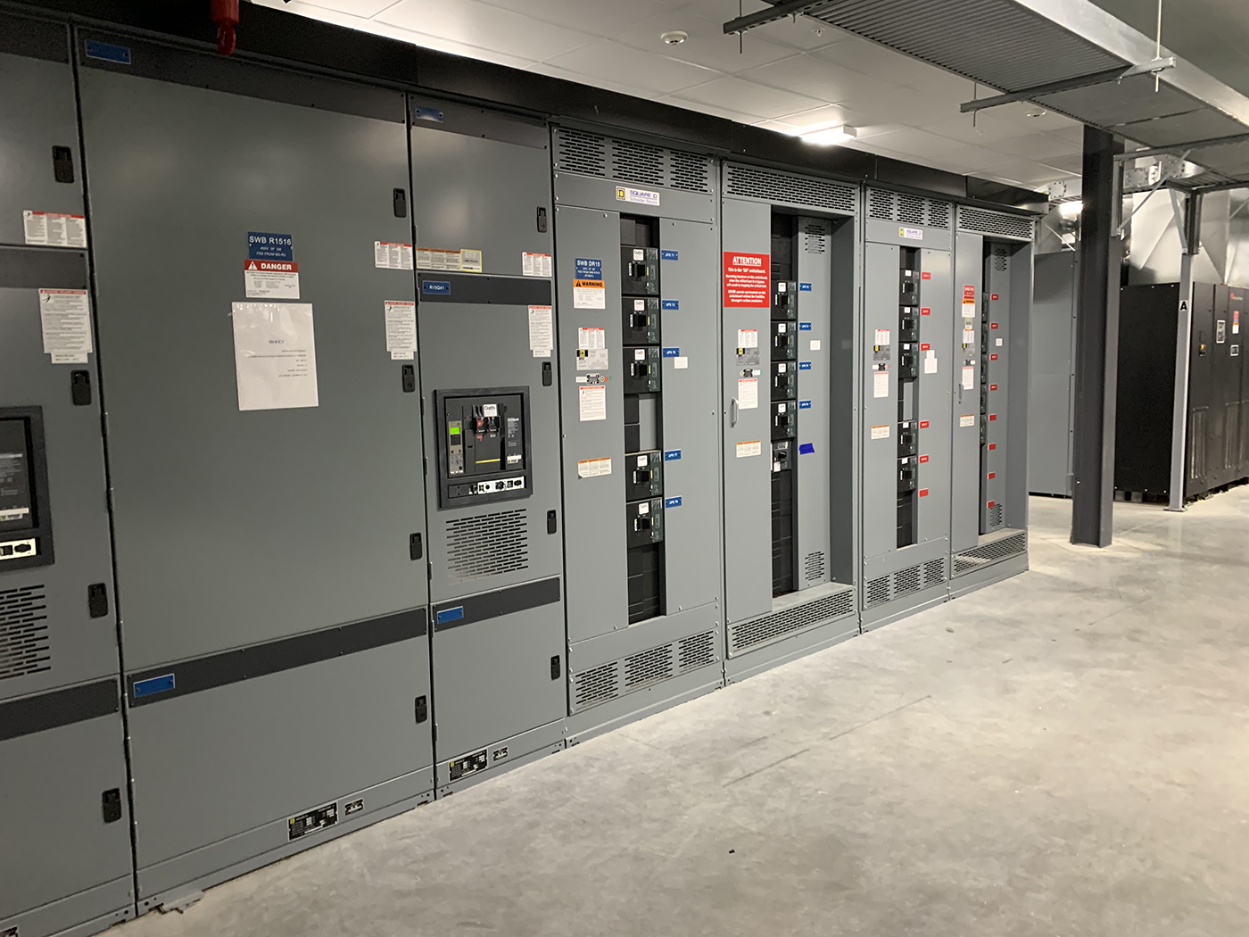 A row of large, grey industrial electrical panels with various buttons, switches, warning labels, and vents is installed along a concrete wall in a clean, modern utility room with a concrete floor.