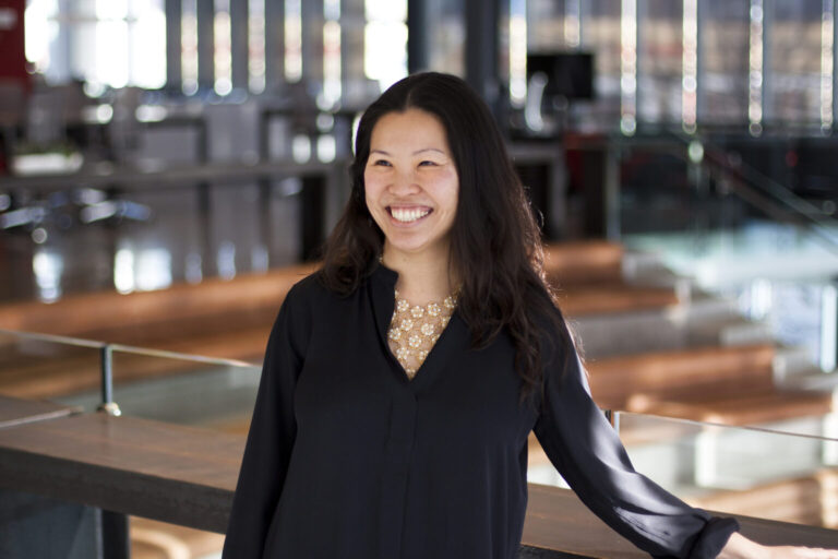 A woman with long dark hair, wearing a black blouse with a gold necklace, stands smiling in a modern, sunlit office space with glass railings and blurred desks in the background.