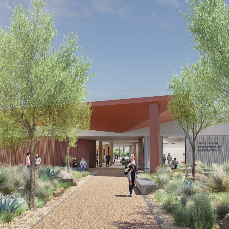 A modern building with a red angular roof, surrounded by desert landscaping and trees. Several people are walking or sitting along a wide, paved path leading to the entrance under a partly cloudy sky.