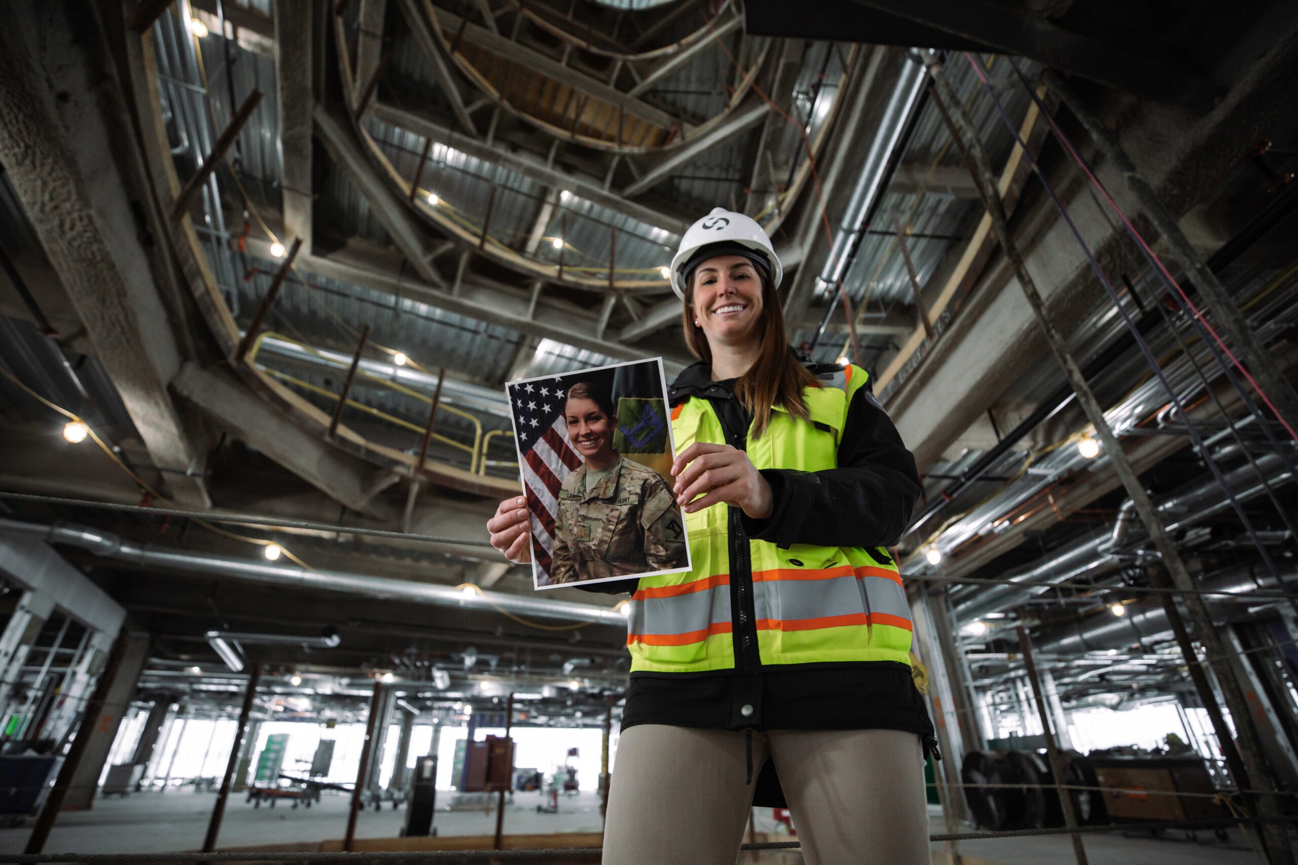 A woman wearing a hard hat and safety vest stands in a large, unfinished building and smiles while holding a photo of a person in military uniform in front of an American flag.