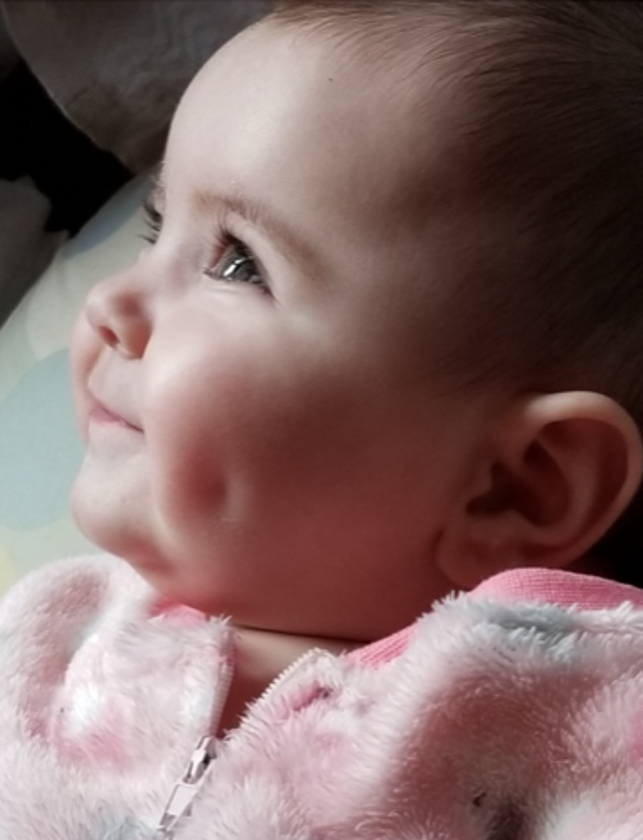 A close-up of a baby with dimples, smiling and looking upward. The baby is wearing a soft, pink, fuzzy outfit and appears content and happy.