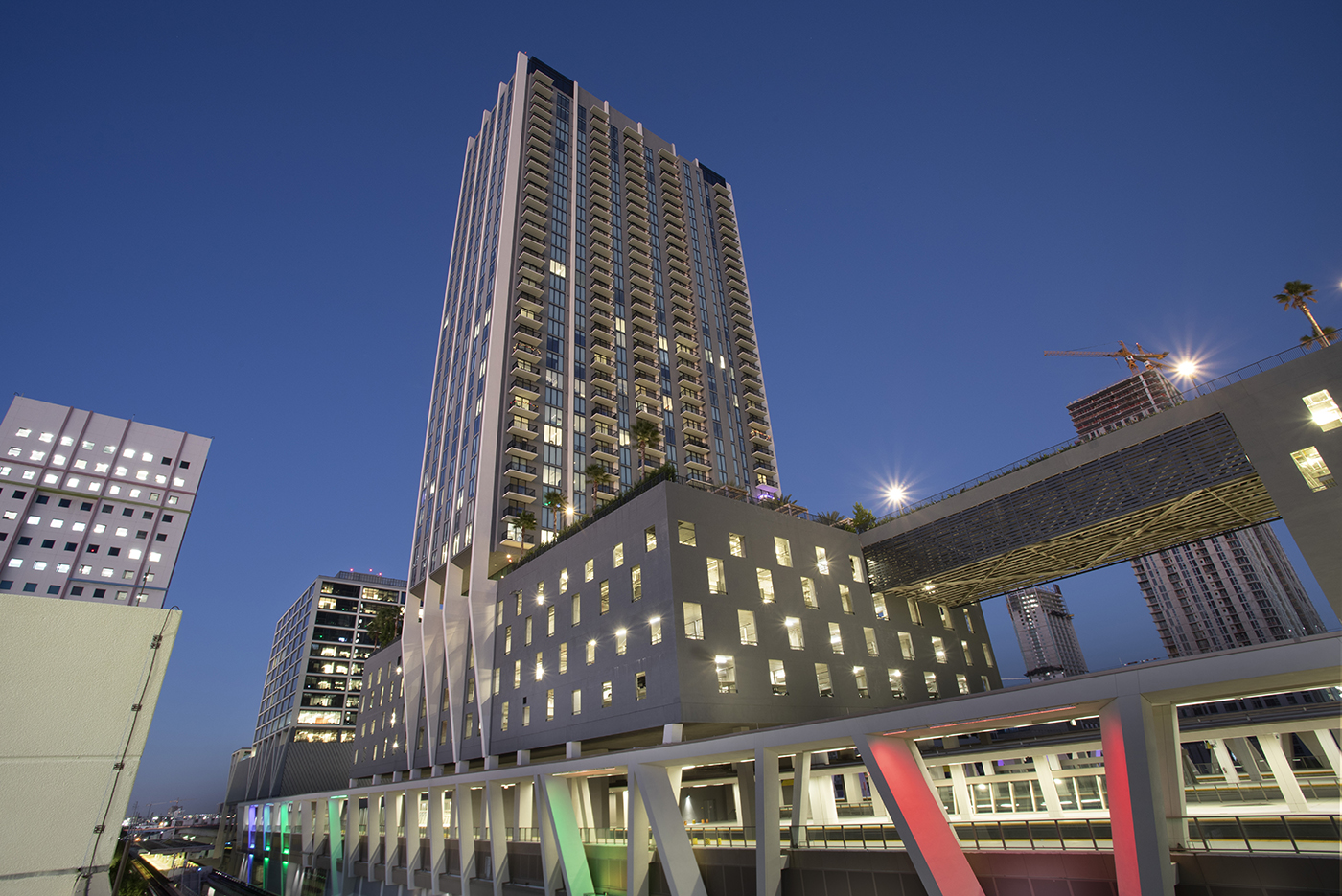 Tall modern high-rise building with illuminated windows and balconies at dusk, with colorful lights on a lower structure in the foreground and other city buildings in the background.