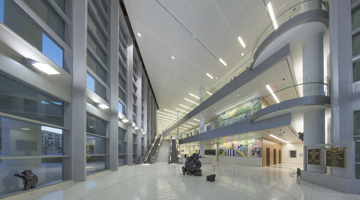 Spacious, modern building interior with high ceilings, large windows, bright lighting, multiple floors, an escalator, abstract sculptures, and colorful artwork on the wall. The floors are shiny and reflective.