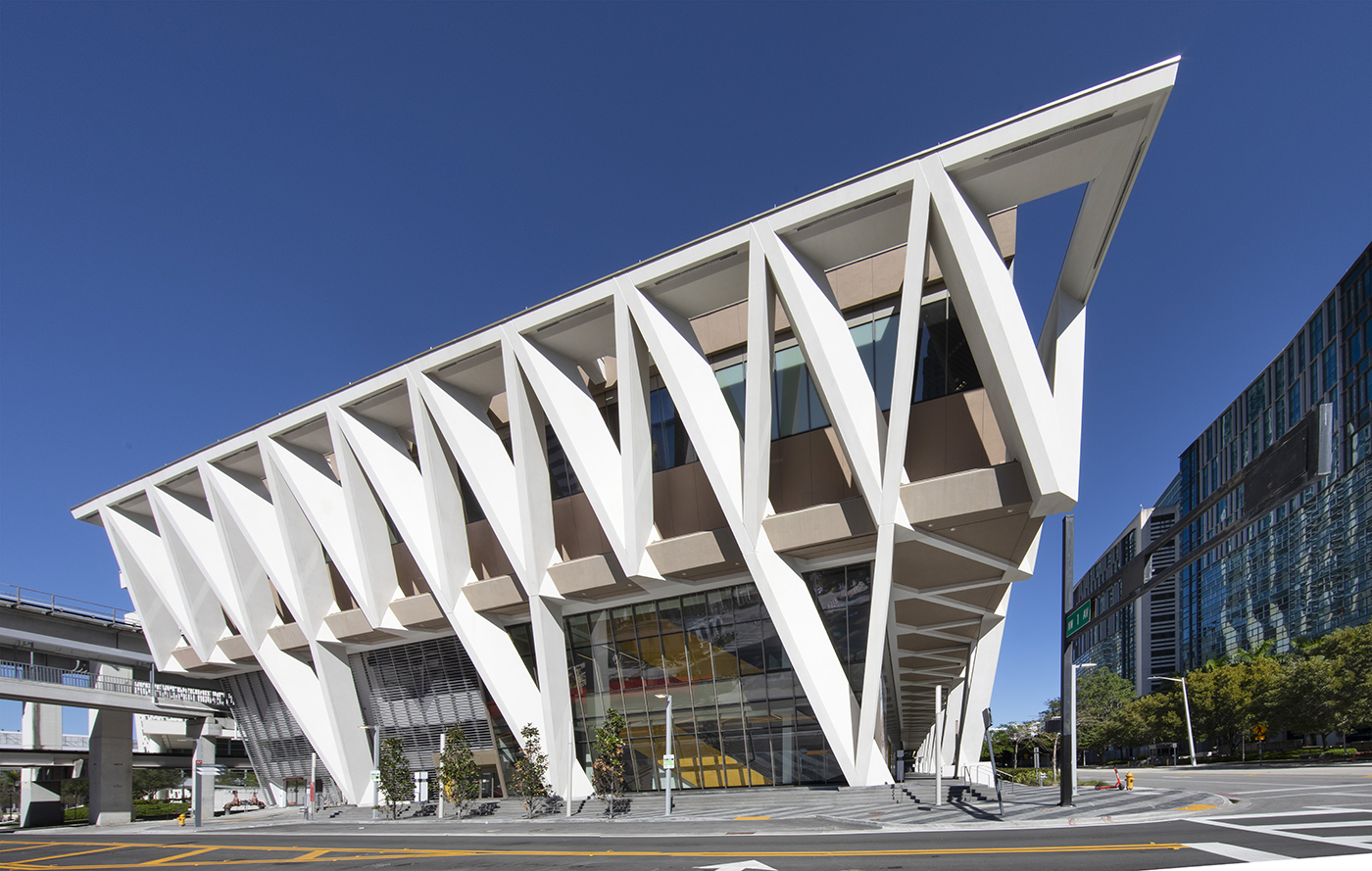 A modern building with dramatic white V-shaped supports and large windows, set against a clear blue sky, with nearby city infrastructure, trees, and roads visible around it.