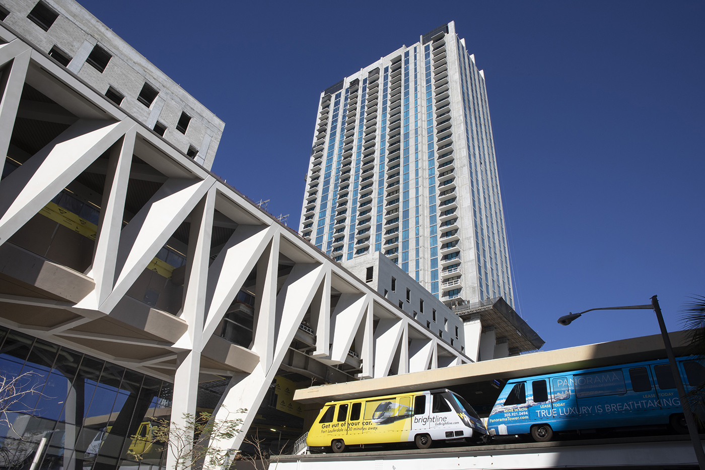 Two colorful trains travel on elevated tracks in front of a modern high-rise building with geometric architecture, under a clear blue sky.