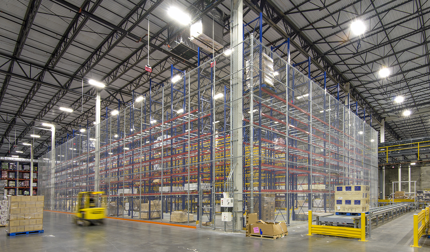 A spacious warehouse interior with tall, empty metal shelving racks, bright overhead lighting, and a yellow forklift moving pallets near boxed goods stacked on the floor. The area is organized and clean, with high ceilings and safety barriers.