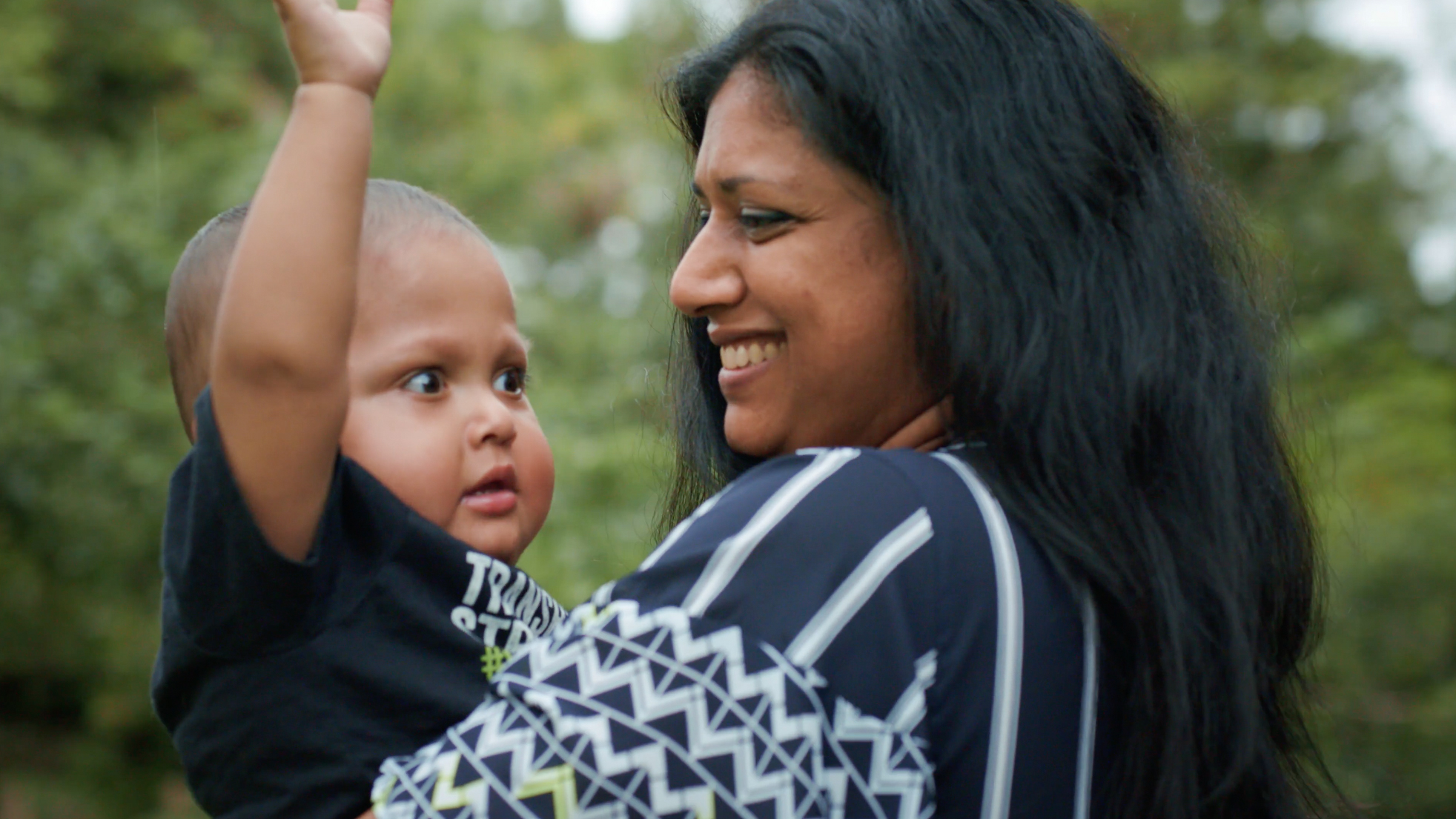 A woman with long dark hair smiles while holding a young child who looks up with one arm raised. They are outdoors with greenery blurred in the background.