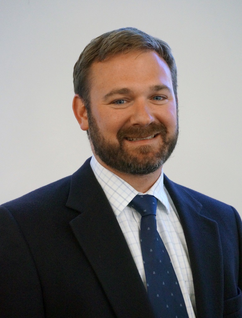 A man with short brown hair and a beard, wearing a dark suit jacket, white checkered shirt, and blue tie, smiles in front of a plain light background.