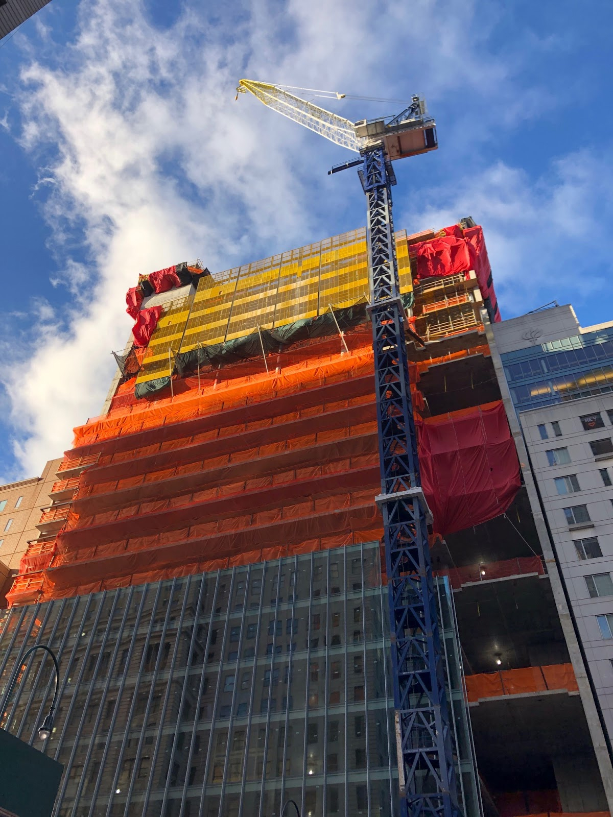 A high-rise building under construction, covered in orange and yellow safety netting, with a tall blue crane positioned in front against a blue sky with clouds.