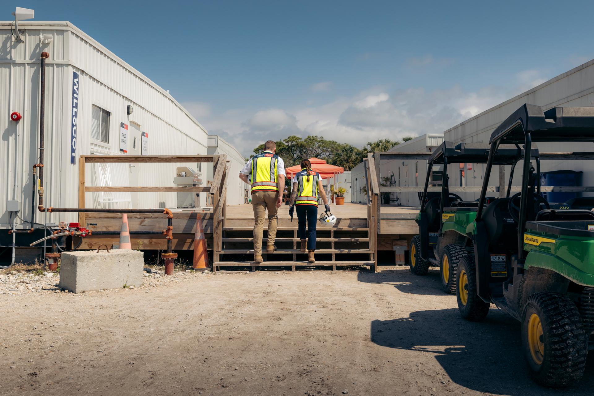 Two construction workers wearing safety vests and hard hats walk up wooden steps between portable site office buildings, highlighting rewarding careers in construction, with utility vehicles parked nearby on a sunny day.