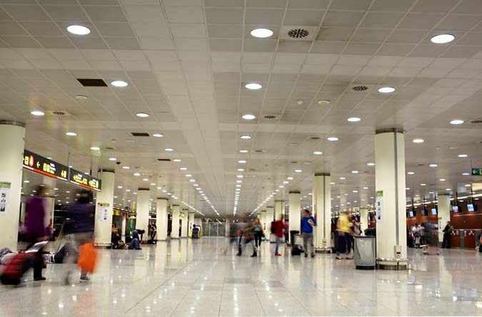 A spacious airport terminal interior with a shiny floor, bright ceiling lights, information boards, and groups of people standing or moving with luggage.