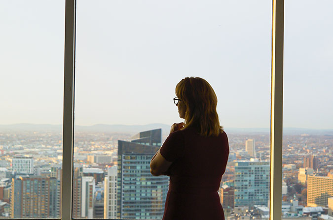 A woman with glasses stands by a large window, looking out over a cityscape with tall buildings and distant hills in the background.