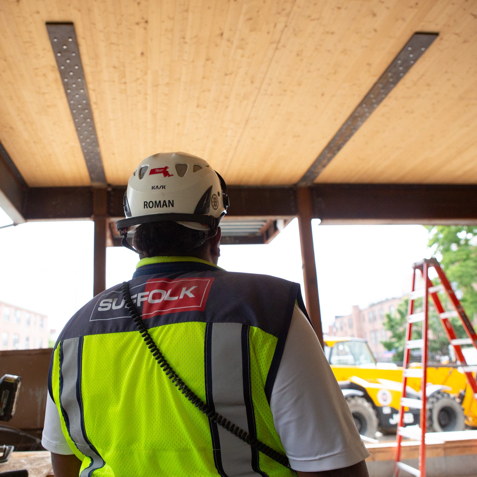 A construction worker in a yellow safety vest and white hard hat stands indoors, facing away. A red ladder and construction equipment are visible outside the building.
