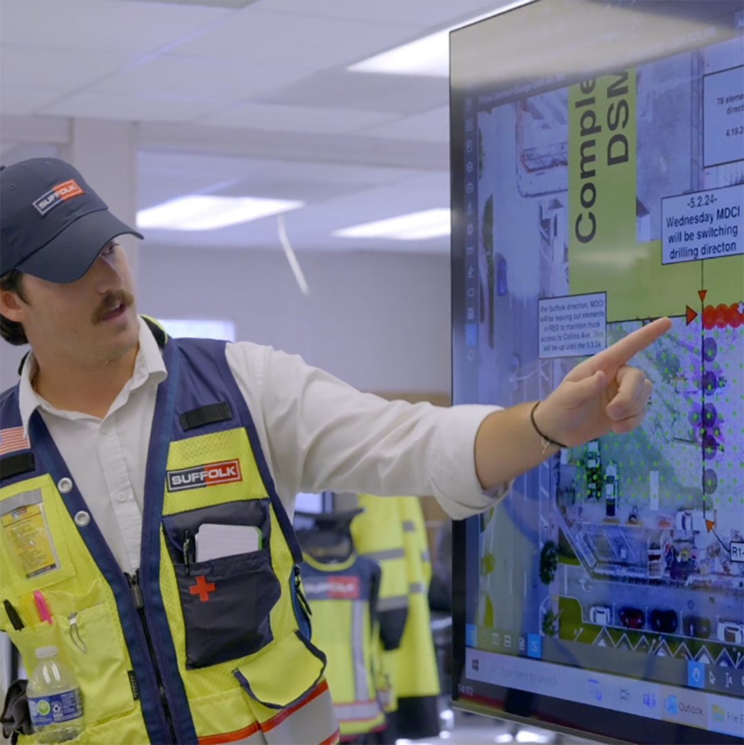 A construction worker in a safety vest and cap points at data on a large touchscreen display in an office setting, explaining information related to a project.