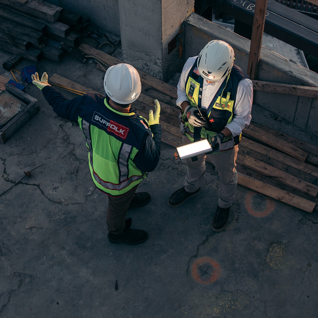 Two construction workers in safety gear and helmets stand on a concrete floor at a construction site, discussing something while one holds a tablet. Wooden planks and construction materials are in the background.