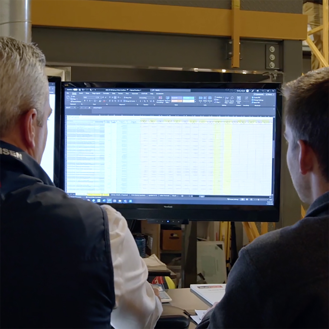 Two people viewed from behind look at a computer monitor displaying a large spreadsheet with rows and columns of data in an office or industrial setting.