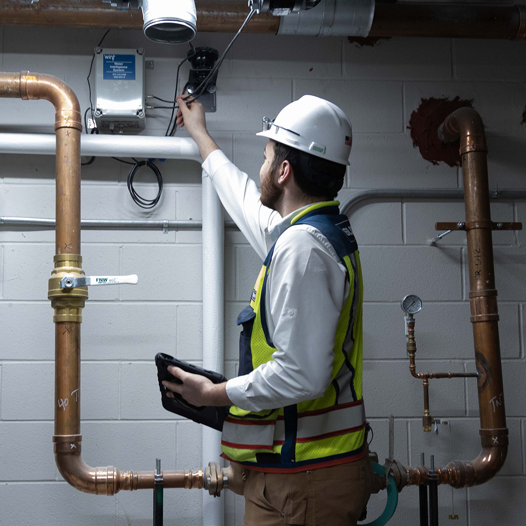 A construction worker in a hard hat and safety vest adjusts a control box mounted on a wall. He stands among copper pipes and holds a tablet, working in an industrial or utility room.
