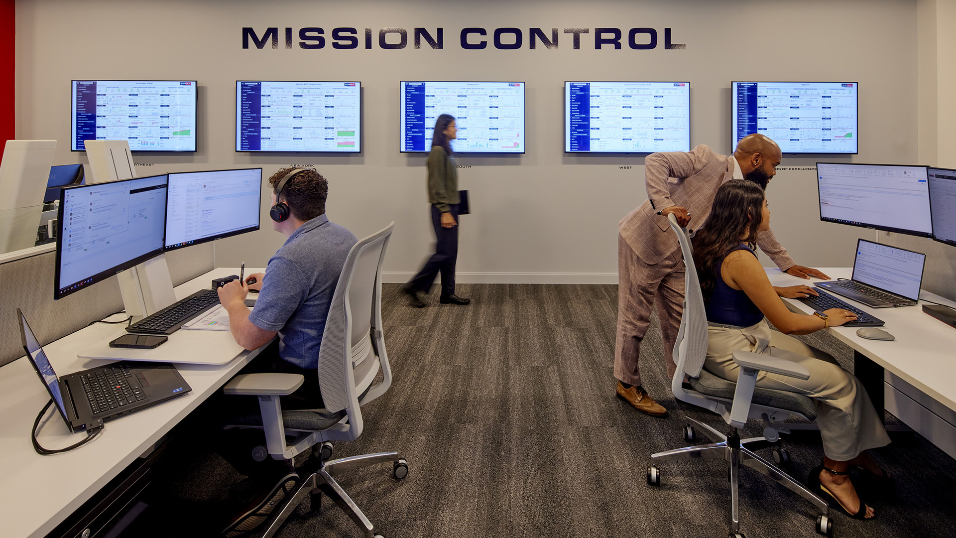 Several people work at computer stations in a modern control room labeled Mission Control, with large screens displaying data on the wall and one person walking by in the background.