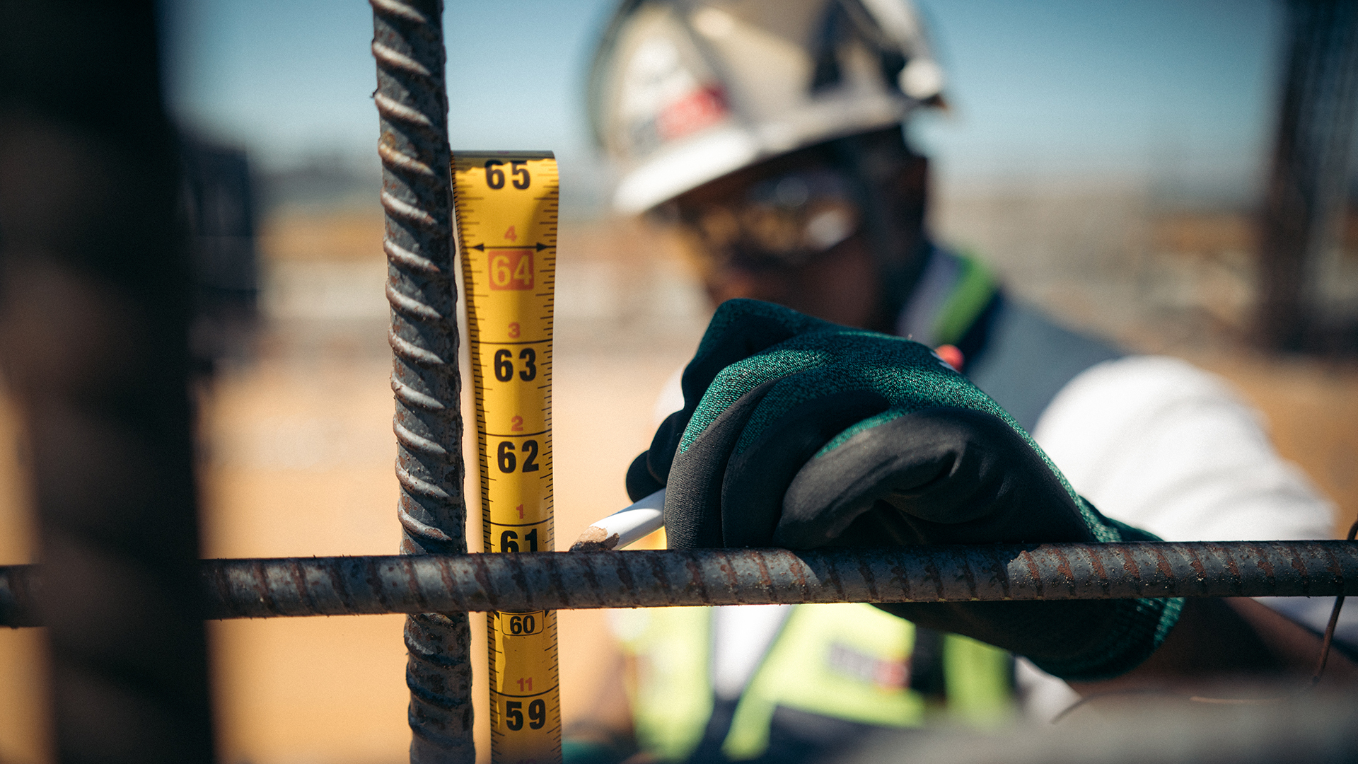 A construction worker wearing gloves and a helmet measures a steel rod with a yellow tape measure and marks it with a pencil. The focus is on the measuring tape and the worker’s gloved hand.