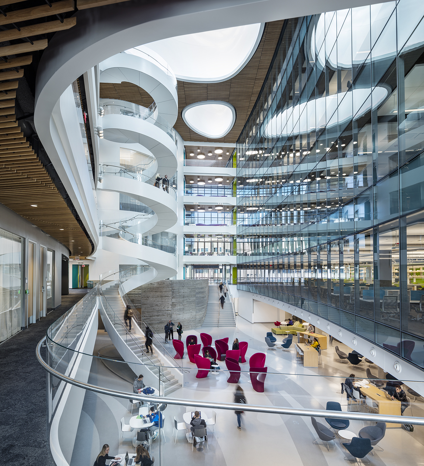 A modern, spacious atrium in an office building with a spiral staircase, large windows, and people working or walking. Red chairs and open seating areas are arranged on light-colored floors under curving architectural features.