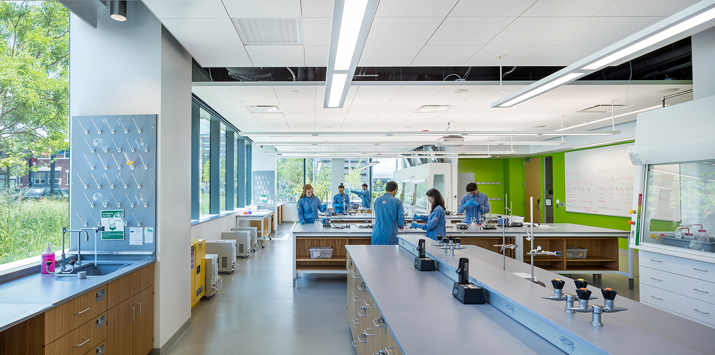 A modern science laboratory with large windows, several people in blue lab coats working at benches, lab equipment on counters, and bright lighting.