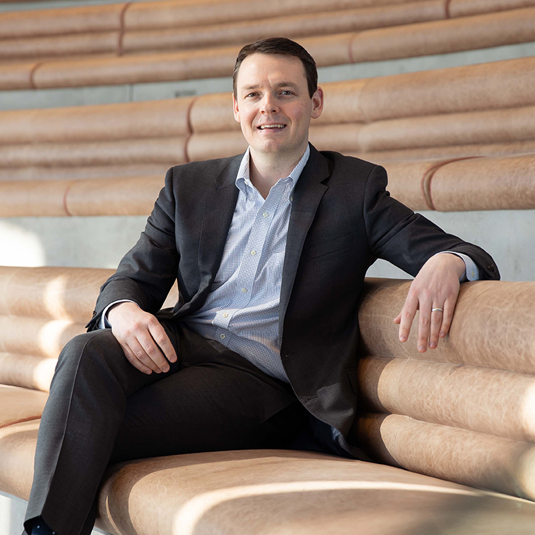 A man in a dark suit and light shirt sits relaxed on tan, curved, tiered seating, smiling at the camera with one arm resting on the back of the seat. The setting appears modern and well-lit.
