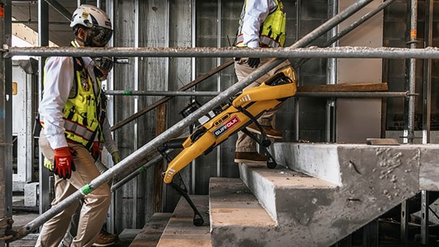 A yellow robotic dog walks up concrete stairs at a construction site while two workers in safety gear and helmets observe. Scaffolding and unfinished walls are visible in the background.