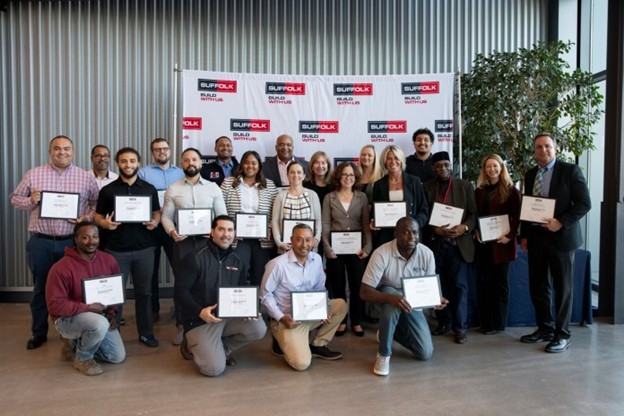 A diverse group of people poses indoors, holding certificates in front of a Suffolk Build With Us banner, celebrating an achievement or graduation.