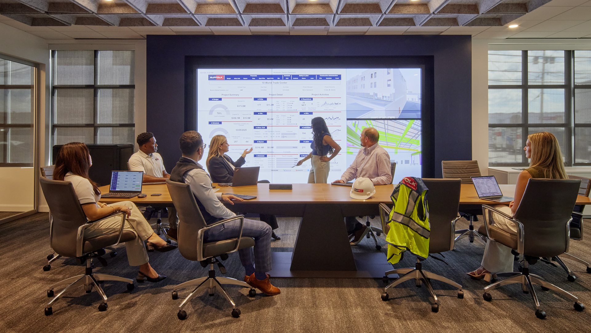 Six people in business attire sit around a conference table in a modern meeting room, viewing and discussing data on a large digital screen; laptops, hard hat, and safety vest are on the table.