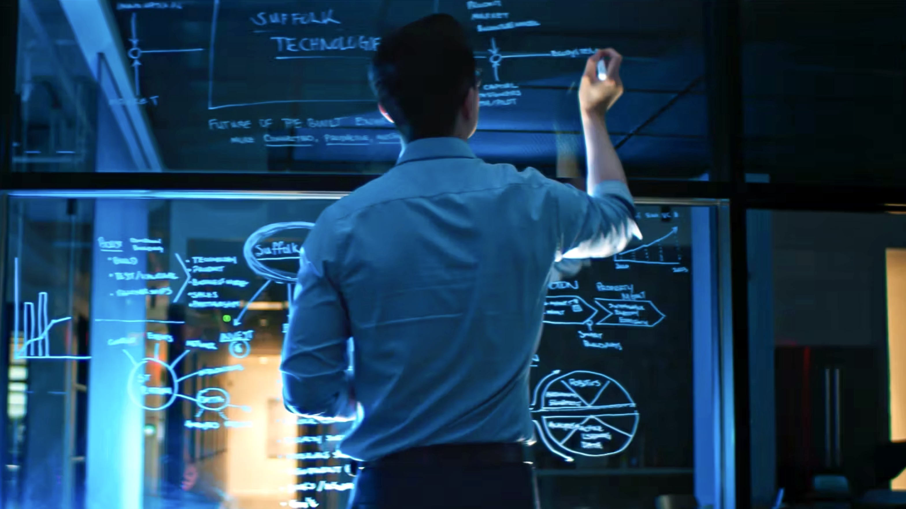 A person in a blue shirt writes on a large transparent glass board covered with diagrams, charts, and notes in a modern, dimly lit office setting. Blue light illuminates the scene.