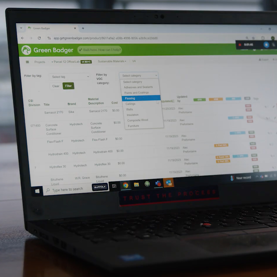 A laptop screen displays the Green Badger app open to a project dashboard, showing data tables and filters for sustainable materials. The mouse pointer hovers over a dropdown menu with categories.