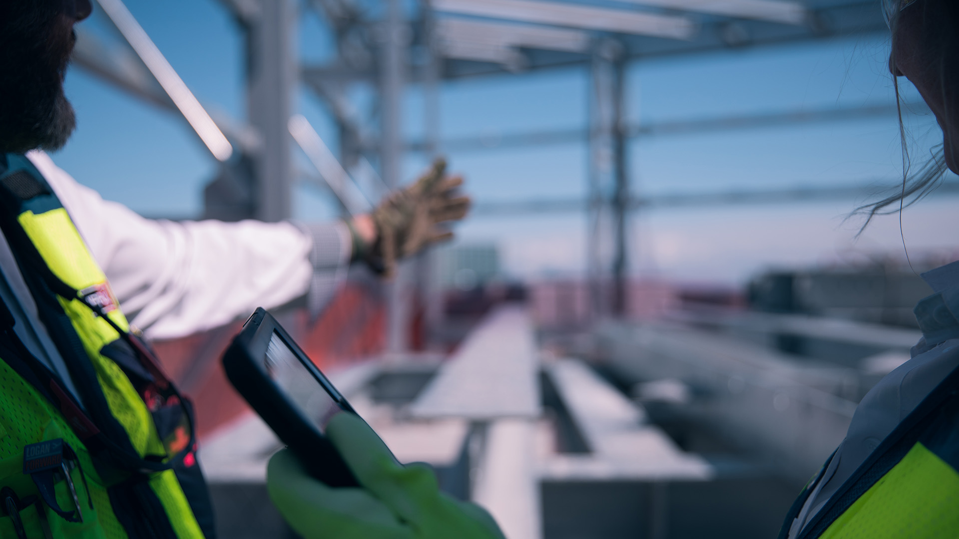 A construction worker wearing a safety vest and gloves holds a smartphone, gesturing toward steel beams at a building site under a clear blue sky.