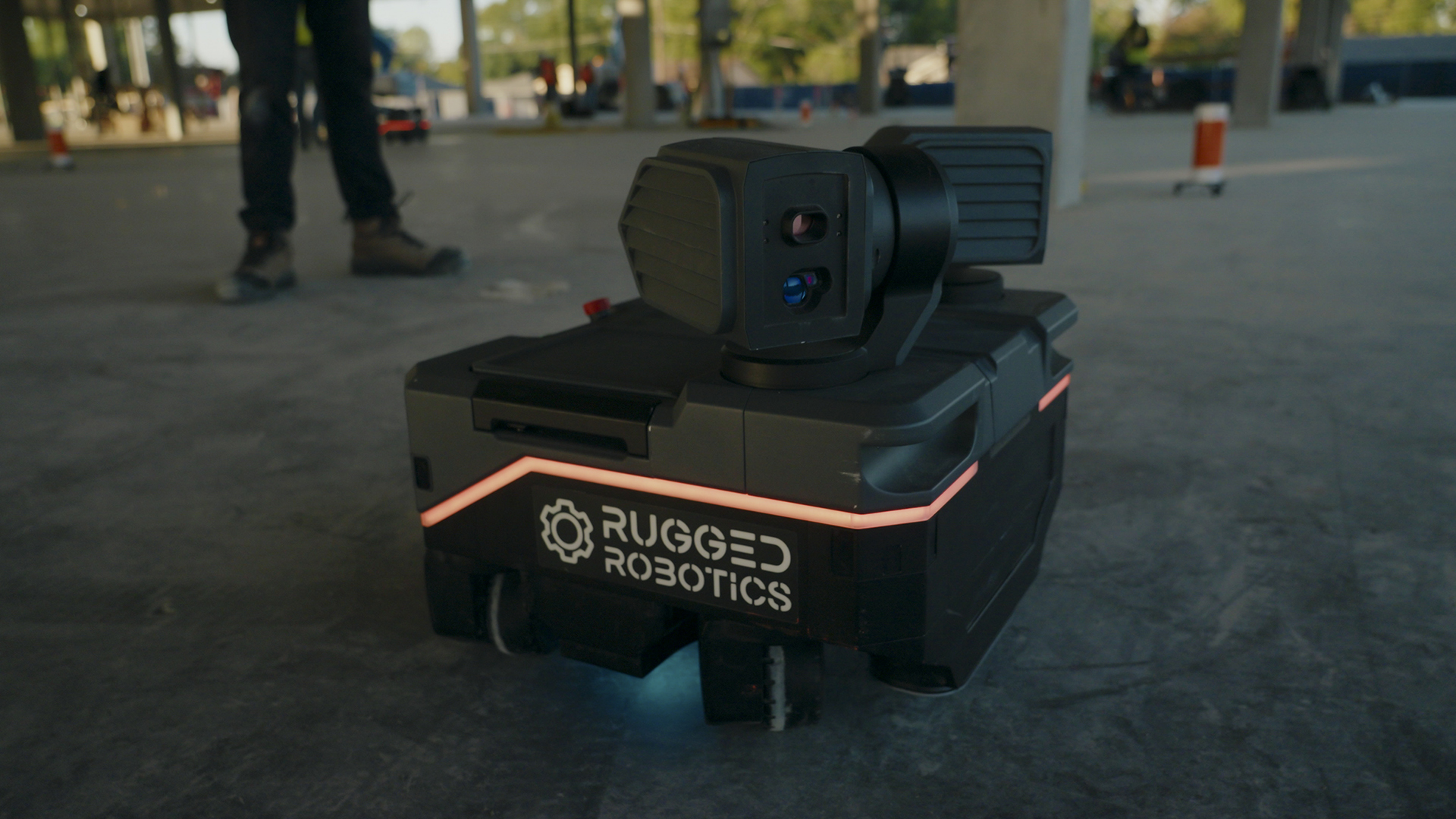 A small, box-shaped robot labeled Rugged Robotics sits on a concrete floor at a construction site, with a person’s legs and unfinished building structure visible in the background.