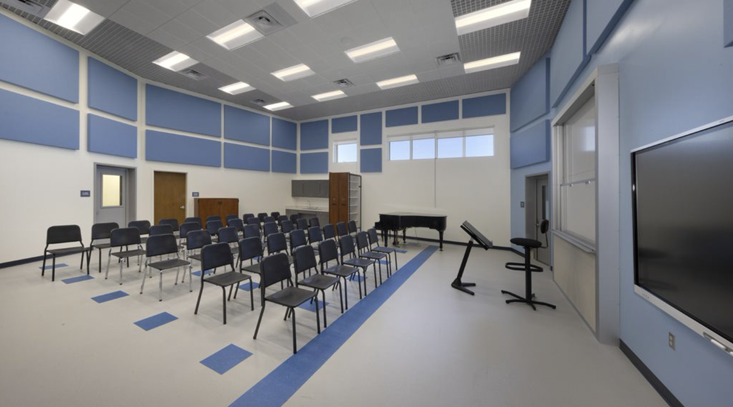 A modern music classroom with rows of black chairs facing a piano, music stands, and a large wall-mounted monitor. The room has blue acoustic panels, bright lighting, and windows near the ceiling.