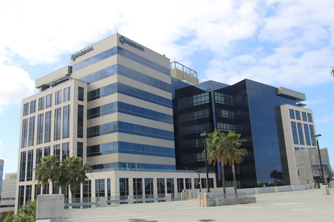 A modern office building with glass and beige panels, multiple stories, and palm trees in front. The sky is partly cloudy, and the scene includes a mostly empty parking area.