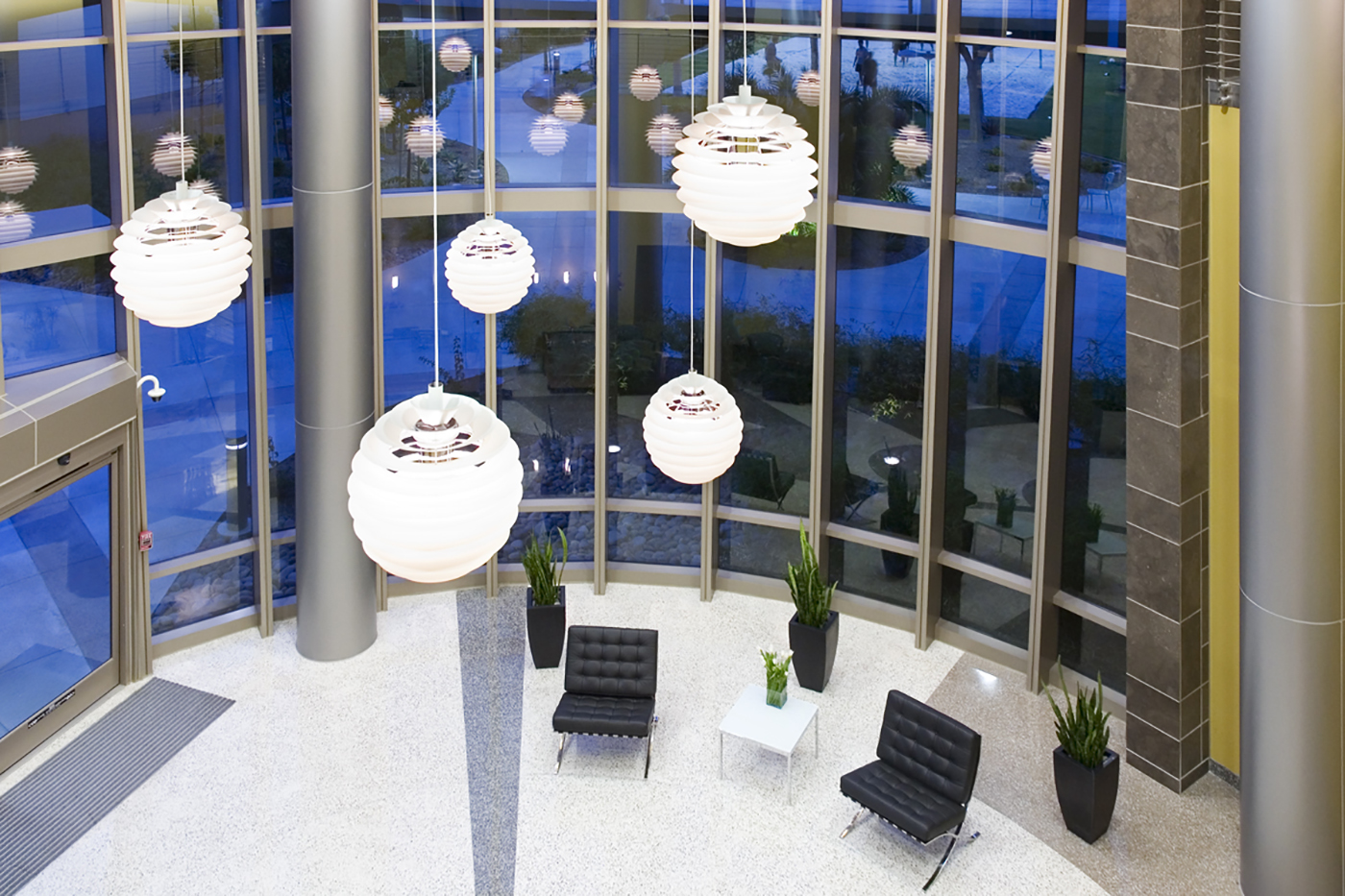 Modern lobby with large round pendant lights, floor-to-ceiling windows, black chairs around white tables, potted plants, and polished stone flooring. Blue light from outside contrasts with interior lighting.