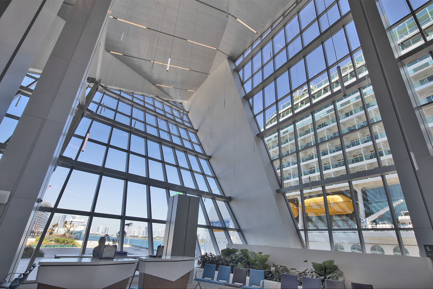 A spacious, modern atrium with tall, slanted glass walls and ceiling, blue chairs, indoor plants, and a view of a docked cruise ship and city buildings through the windows.