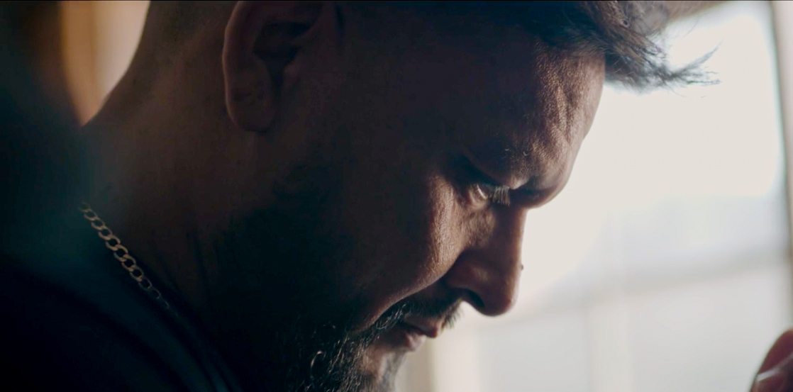 A close-up side view of a man with a beard and short hair, wearing a chain necklace, looking downwards with a serious or focused expression. Sunlight softly illuminates his face from the background.