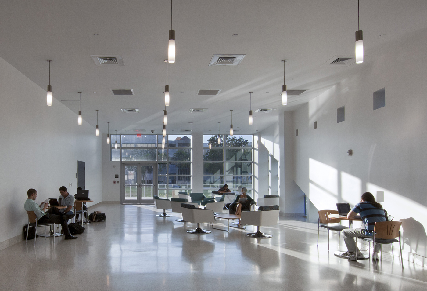 A modern, spacious lounge area with large windows, white walls, and overhead lights. Several people are sitting on chairs or working at tables, some using laptops, while sunlight streams in through the windows.