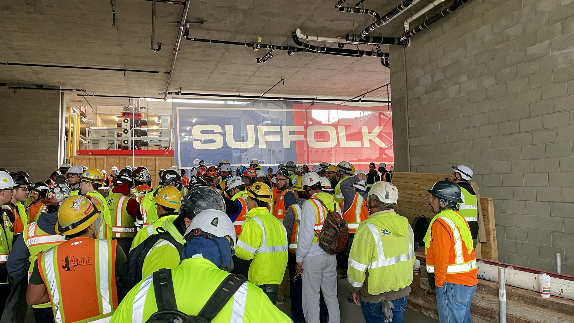 A large group of construction workers in safety vests and helmets gather indoors near a wall of cinder blocks. In the background, a window displays a large SUFFOLK sign in red and white letters.