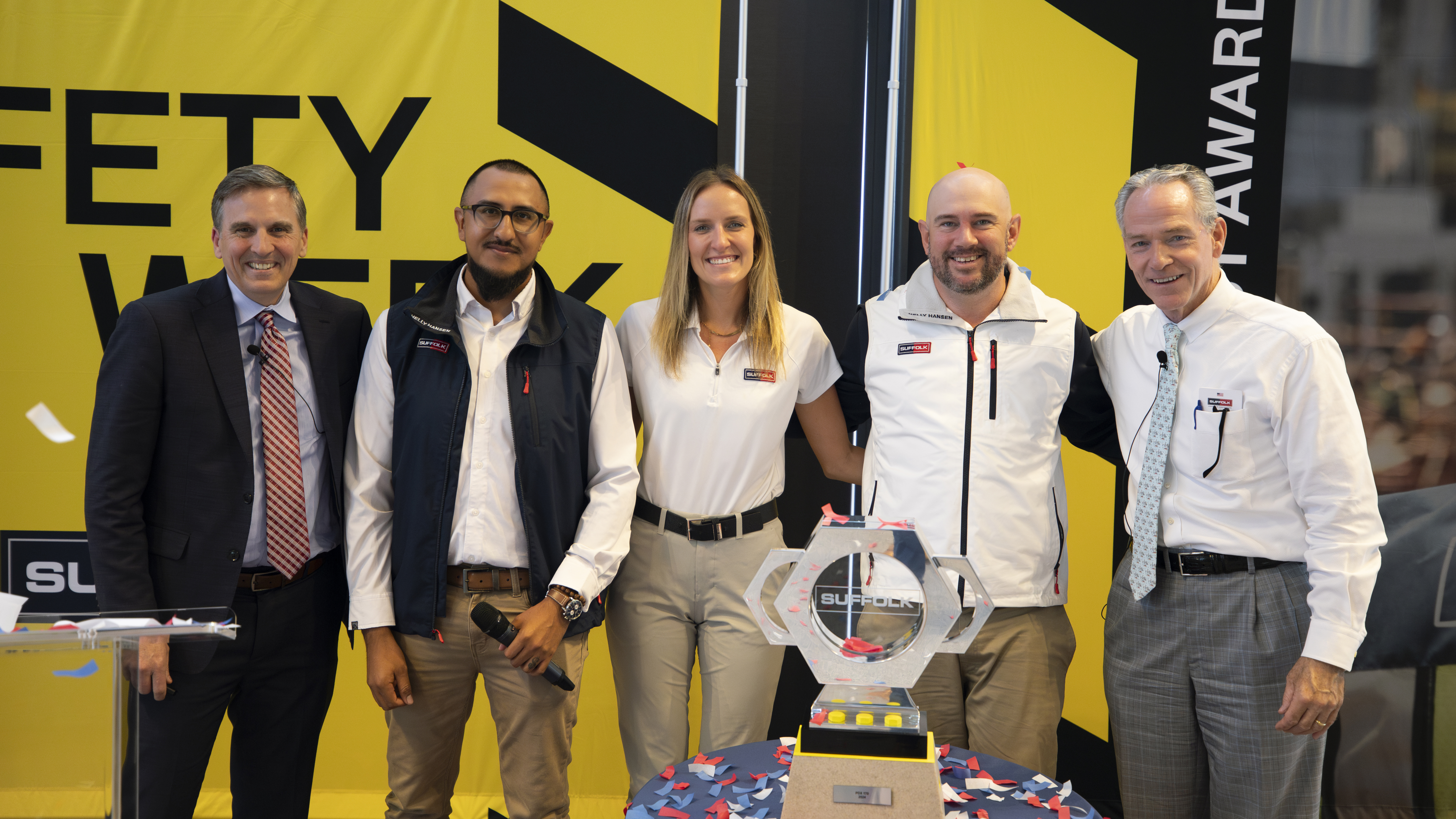 Five people stand smiling in front of a yellow Safety Week backdrop, with a trophy on a table in front of them. Blue and red confetti is scattered around the trophy.