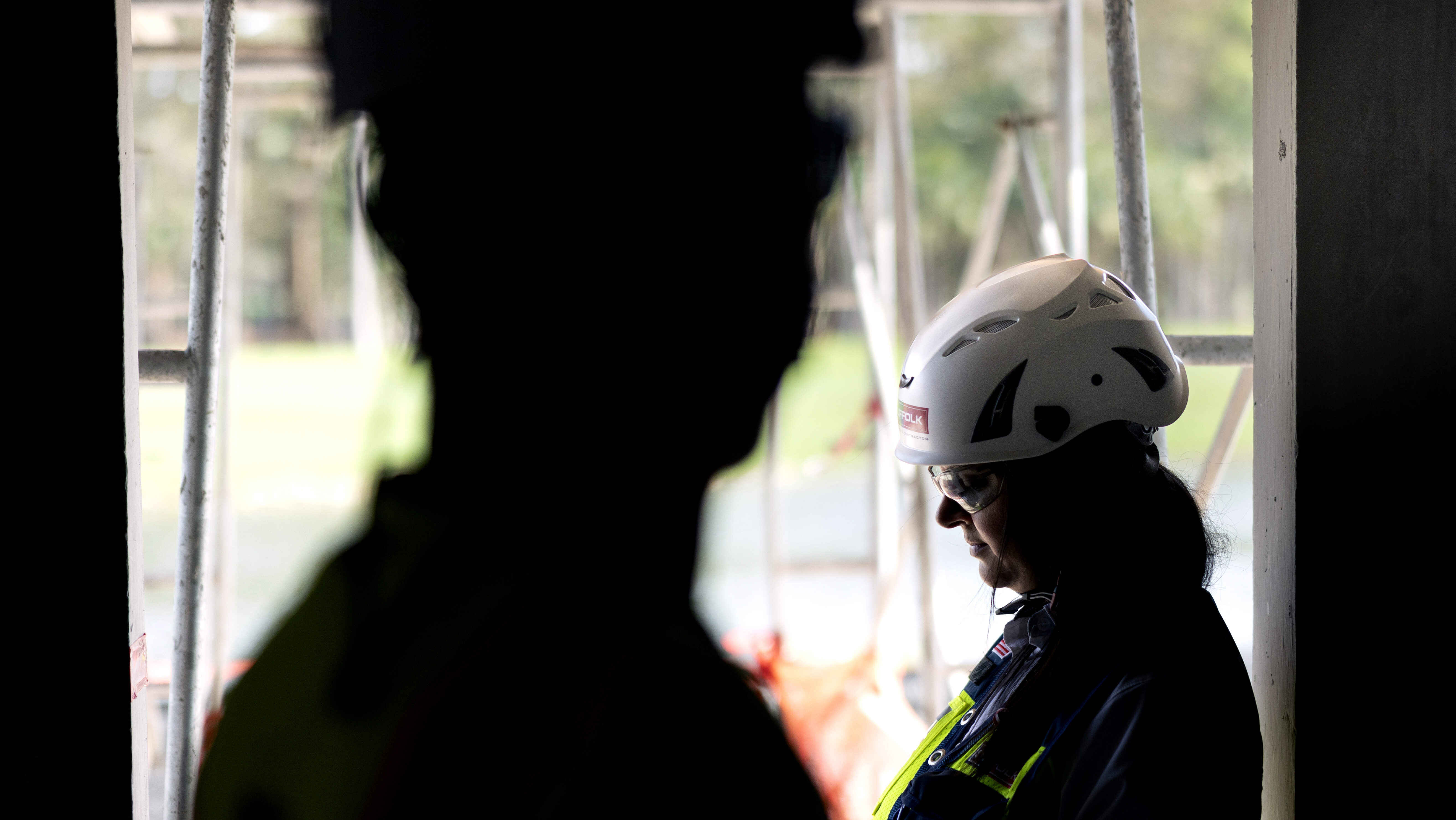 A person wearing a white safety helmet and dark clothing stands in profile, looking down, with another person blurred in the foreground. Outdoor scaffolding and greenery are visible in the background.