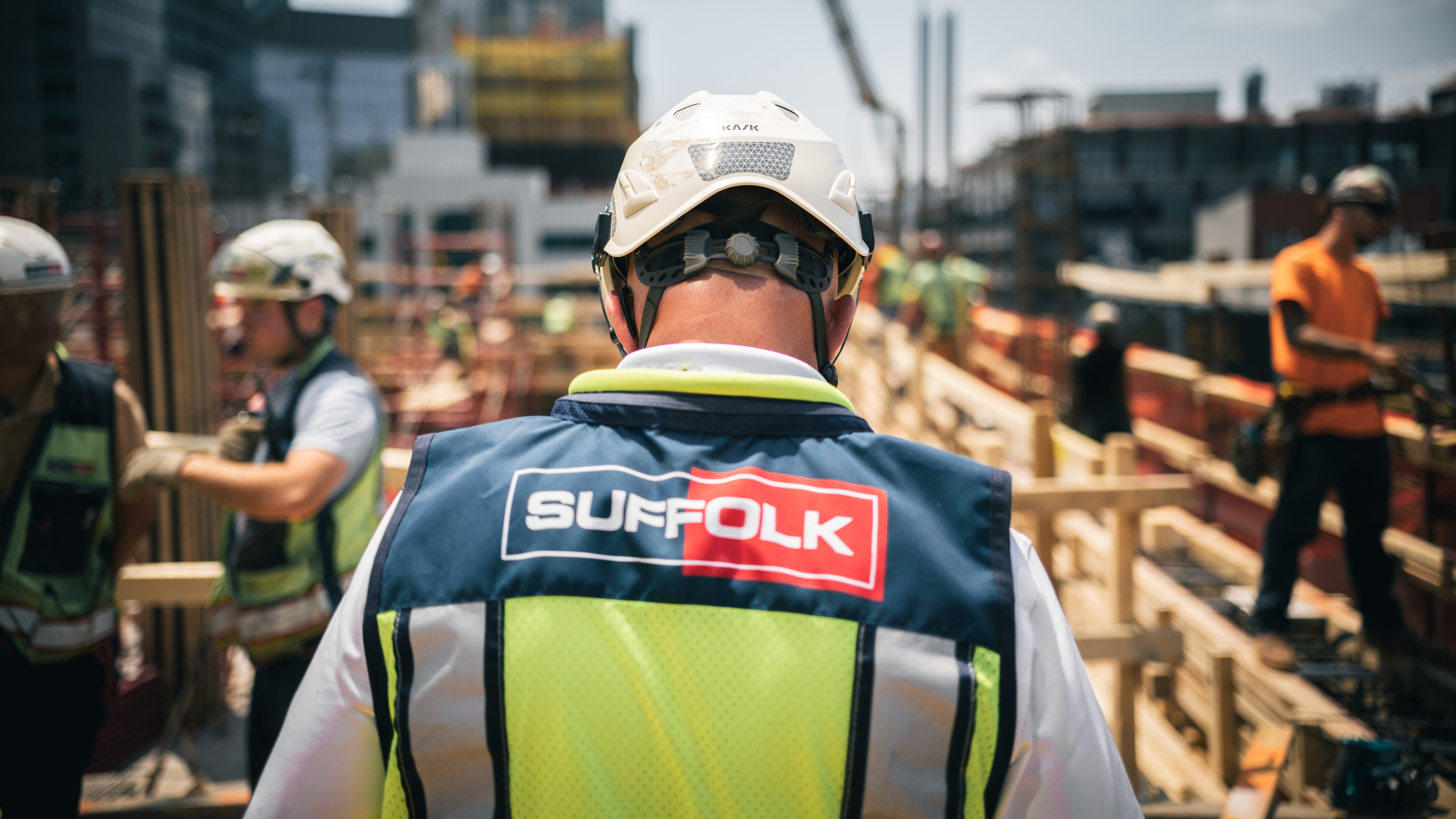 A construction worker wearing a helmet and a vest labeled “Suffolk” stands on an active construction site, with other workers and building materials visible in the background.
