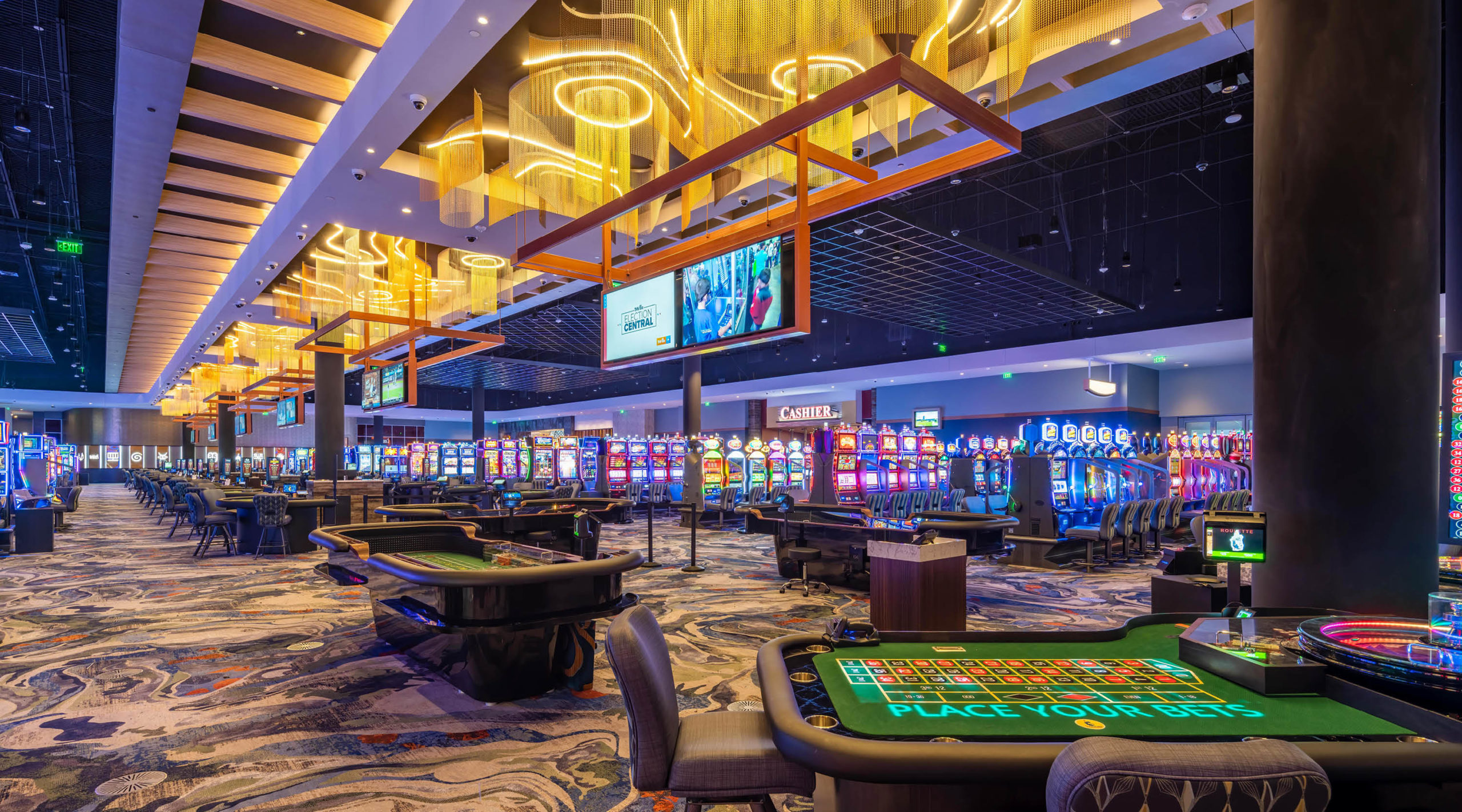 A brightly lit casino interior featuring rows of slot machines, electronic screens, and gaming tables for roulette and cards on a patterned carpet under modern golden ceiling lights.
