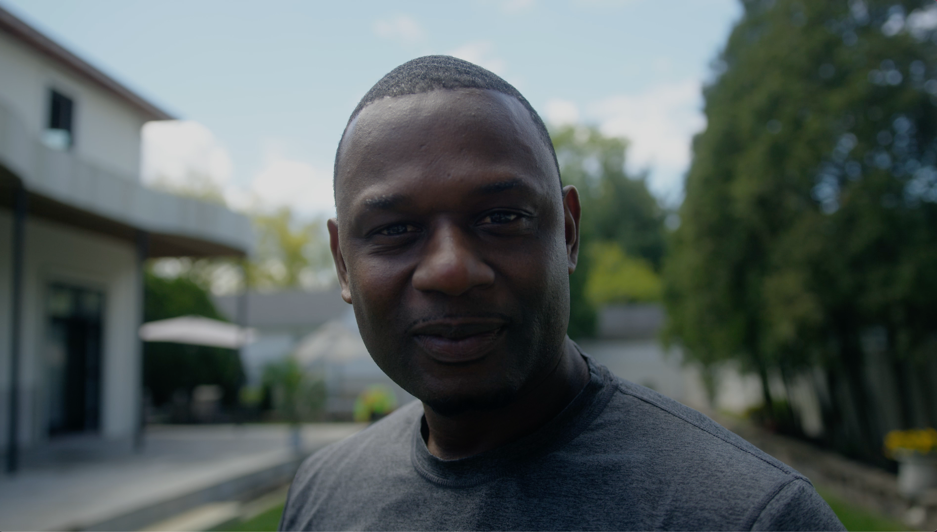 A man wearing a dark gray t-shirt stands outdoors, smiling slightly at the camera, with trees, a house, and a patio area blurred in the background.