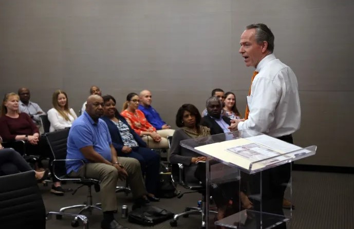 A man in a white shirt speaks at a clear podium to a seated group of adults in a conference room, with attendees listening attentively.